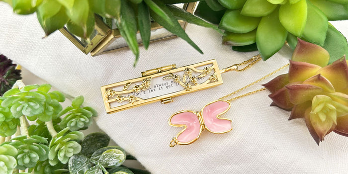 Gold Flowered Vines Fortune Locket and Gold Fortune Cookie Locket (Blush) surrounded by plants.